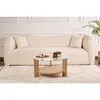 A cream-colored sofa and a round coffee table with a glass top and wooden base are placed on a fluffy white rug in a well-lit room. Produktbild-Vorschau