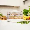 A food dehydrator filled with food is placed on a white countertop in a kitchen setting. Other food items like bananas, strawberries, mushrooms, and nuts are also visible. Náhľad obrázku produktu