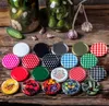 A collection of colorful jar lids arranged on a wooden surface, with jars of pickles and garlic in the background. Podgląd zdjęcia produktu