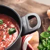 A close-up shot of a pot handle with a logo, filled with tomato sauce and garnished with basil leaves, resting on a wooden surface with a striped cloth. Náhled obrázku produktu