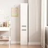 A tall, narrow white cabinet with two doors stands next to a wall with some artwork and a desk with books. Podgląd zdjęcia produktu
