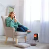 A woman sits in a chair wrapped in a blanket, holding a mug and talking on the phone, with a small electric heater in the background. Anteprima dell’immagine prodotto
