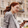 A woman with red hair is wearing earbuds and looking at her phone while walking outside. Náhled obrázku produktu