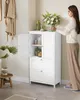 A woman is placing a vase of flowers on top of a white bathroom cabinet. The cabinet has two doors and two drawers, and is filled with toiletries and towels. A bathrobe hangs on the wall behind it, and a plant sits on a stool to the left. Produktbild-Vorschau