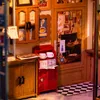 A miniature bistro scene with wooden cabinets, a red cabinet with cups and saucers, and a checkered floor. Voir les images du produit