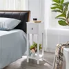 A white nightstand with a drawer and shelves is placed next to a bed. A glass of dark liquid and a book are on top of the nightstand. A vase with flowers is on the lower shelf. A plaid scarf is draped over a white stool in the foreground. Produktbild-Vorschau
