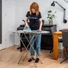 A woman is ironing clothes on a foldable ironing board in a room with a piano and dining table. Podgląd zdjęcia produktu