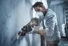 A man wearing safety gear is using a reciprocating saw to cut a pipe against a concrete wall. Produktbild-Vorschau
