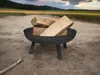 A black metal fire pit filled with logs sits on a dirt path, with a field and trees in the background under a cloudy sky. Voir les images du produit