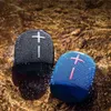 Two portable speakers are submerged in shallow water, with water splashing around them. Produktbild-Vorschau