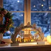 A wooden Christmas decoration with lights sits on a windowsill in front of a snowy window. Náhľad obrázku produktu