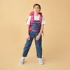 A young girl wearing a backpack and overalls stands against a plain background. Anteprima dell’immagine prodotto