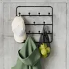 A black metal wall-mounted rack with hooks, holding two baseball caps, a green hooded garment, a mesh bag with yellow fruits, against a concrete wall. Produktbild-Vorschau