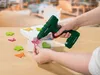 A person is using a green and red cordless hot glue gun to attach a pink butterfly decoration to a white box. Several other colorful butterfly and clover-shaped decorations are scattered on the wooden surface. Produktbild-Vorschau