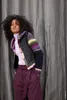 A woman leans against a gray curtain, wearing a colorful knitted cardigan. Produktbild-Vorschau