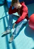 A young man wearing a red hoodie and light blue jeans sits on a blue floor, leaning his head on his hand. He is wearing Vans checkerboard slip-on shoes. A large red exercise ball is visible to his right. Voir les images du produit