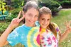 Two young girls are posing for a photo outdoors, smiling and making peace signs. They are wearing colorful tie-dye t-shirts. Produktbild-Vorschau