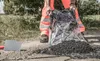 A person in high-visibility orange work clothes is pouring black asphalt from a clear plastic bag onto the ground next to a road or path. A trowel is visible on the left. Produktbild-Vorschau