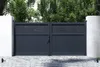 A dark gray double gate with decorative cutouts at the top, set between two white pillars. The gate is in front of a backdrop of green trees and is situated on a paved driveway. Produktbild-Vorschau