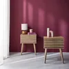 Two Scandinavian-style bedside tables with slatted wood fronts are placed against a deep red wall. One table has a lamp, books, and a small decorative item on top, while the other has a decorative vase. Produktbild-Vorschau