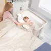 A mother is lying in bed next to a baby in a bedside bassinet. The mother is reaching out to touch the baby. Produktbild-Vorschau