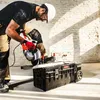A construction worker is using an angle grinder to cut a metal bar, with a Qbrick System tool box in the foreground. Náhled obrázku produktu