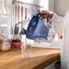 A person is pouring water from a blue filter pitcher into a clear glass on a wooden countertop. Various kitchen utensils hang in the background. Produktbild-Vorschau