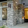 An outdoor wall light fixture is mounted on a stone pillar next to a large window of a modern house. Produktbild-Vorschau