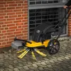 A person is using a yellow and black powered weed brush to clean a paved area next to a brick wall and a window with security bars. Produktbild-Vorschau