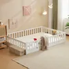 A white wooden children's bed with a mattress and bedding, set up in a room with decorative art on the wall and a floor lamp. Náhľad obrázku produktu