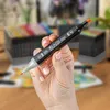 A hand holding a black marker with an orange tip, with stacks of markers in the background. Produktbild-Vorschau