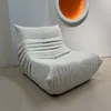 A beige armchair is placed in a room with a white wall and a light-colored floor. An abstract orange art piece is mounted on the wall behind the chair. Produktbild-Vorschau