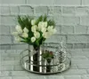 A silver cylindrical vase with white tulips and greenery sits on a mirrored tray, next to a small succulent and a silver pineapple decoration, against a brick wall background. Produktbild-Vorschau