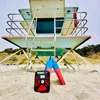 A red and black Eton Quest DAB+ emergency radio sits on a sandy beach in front of a lifeguard tower. Two red and blue fins are propped up behind the radio. Produktbild-Vorschau