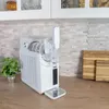 A white slushy maker sits on a wooden countertop next to a potted plant, with kitchen cabinets and a tiled backsplash in the background. Produktbild-Vorschau