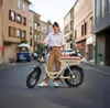 A woman stands next to an electric bicycle on a street in a European town. Produktbild-Vorschau