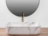 A close-up view of a marble-patterned vessel sink with a gold faucet, set against a plain white wall and a round mirror. Produktbild-Vorschau