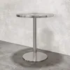 A small, round, silver table with a single pedestal base is shown against a textured gray wall and concrete floor. Produktbild-Vorschau