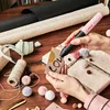 A person is using a pink and black Bosch Gluey hot glue gun to write on a small card, surrounded by craft supplies like twine, pom-poms, and wrapping paper. Produktbild-Vorschau