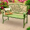 A green metal garden bench with a decorative backrest and a cushion, placed outdoors with flowers in the background. Podgląd zdjęcia produktu