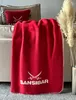 A red blanket with white text and crossed swords logo is draped over a couch, next to a pillow with the same logo. A vase with dried pampas grass is also visible. Produktbild-Vorschau