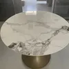 A round dining table with a marble-like ceramic top and a gold pedestal base is shown in a room. Produktbild-Vorschau