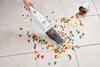 A hand is using a white handheld vacuum cleaner to pick up colorful gummy bears spilled on a tiled floor from a silver cup. Produktbild-Vorschau