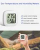A hand holding a small digital thermometer and hygrometer with a blurred background of what appears to be a terrarium. Produktbild-Vorschau