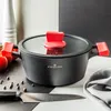A black pot with red handles and lid knob sits on a checkered surface, with tomatoes, peppercorns, and a knife nearby. Podgląd zdjęcia produktu