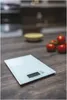 A digital kitchen scale on a wooden countertop with tomatoes and a bowl in the background. Anteprima dell’immagine prodotto