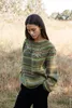 A woman wearing a sweater stands in a field of dry grass and trees. Produktbild-Vorschau