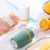 A close-up shot of two small portable vacuum sealers, one white and one green, with a vacuum-sealed bag of chicken in the foreground. Oranges and corn are also visible in the background. Produktbild-Vorschau