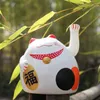 A white Maneki-neko (lucky cat) figurine sits on a wooden post, partially obscured by green bamboo leaves. Produktbild-Vorschau