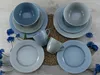 A set of blue and gray ceramic dishes, including plates, bowls, and mugs, arranged on a woven placemat. Produktbild-Vorschau